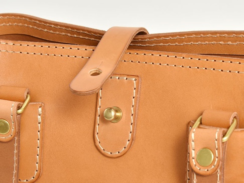 職人手作皮革原子釦托特包，採用天然原色皮革製作，包身縫線細緻，配有金屬原子釦及皮革繫帶，展現簡約質感。