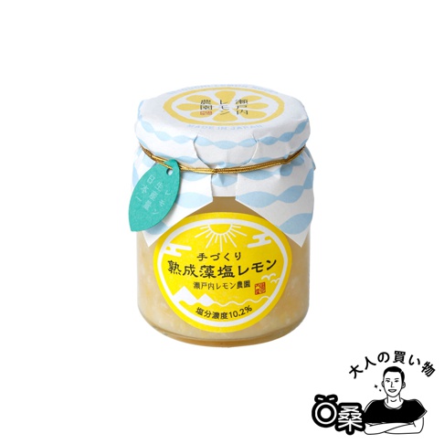 玻璃瓶裝熟成藻鹽漬檸檬，內容物呈現淡黃色，瓶蓋印有藍白色波浪圖案與黃色圓形標籤，標籤上繪有太陽圖案及日文說明，並繫有金色繩子及藍綠色小吊牌。