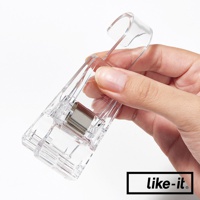 一隻手握著透明塑膠製的雙頭萬用掛式小物夾，夾子上有金屬彈簧，設計簡潔，適合夾取各種小物。