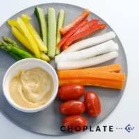 灰色圓形砧板上擺放著豐富的切塊蔬菜，包含蘆筍、黃椒、小黃瓜、紅椒、白蘿蔔、紅蘿蔔，以及一碗醬料和數顆小番茄。