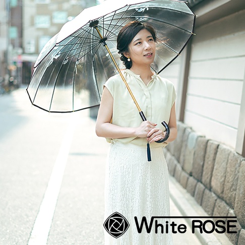 一位女性手持一把透明傘，傘骨為黑色，傘柄為樺木材質，帶有黑色流蘇。她身穿淺黃色無袖上衣和白色蕾絲長裙，站在街道上。