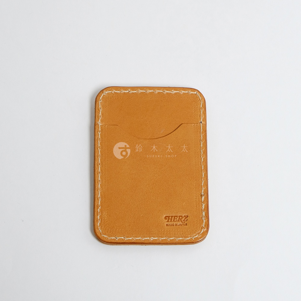 一個 tan 色的皮革名片夾，有清晰的縫線，上方有一個弧形開口，中央印有 SUZUKI SHOP 的標誌和日文，下方印有 HERZ MADE IN JAPAN 的字樣。