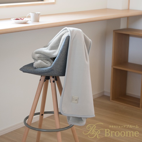 淺灰色絨面保暖毯，一角疊放在灰色布藝高腳椅上，毯子邊緣有淺藍色滾邊，毯子上帶有品牌標籤。