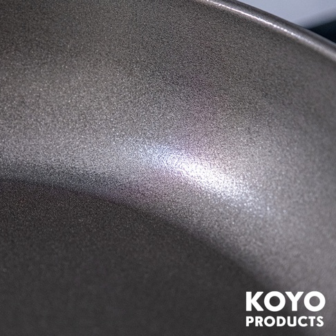 這張近距離特寫照片展示了 KOYO PRODUCTS 輕量壓紋淺型鐵製平底鍋的內鍋表面，金屬表面呈現帶有細緻顆粒感的壓紋設計，深灰色的鍋面在光線照射下泛出微微金屬光澤，展現出材質耐用且具防沾黏功能的質感細節。