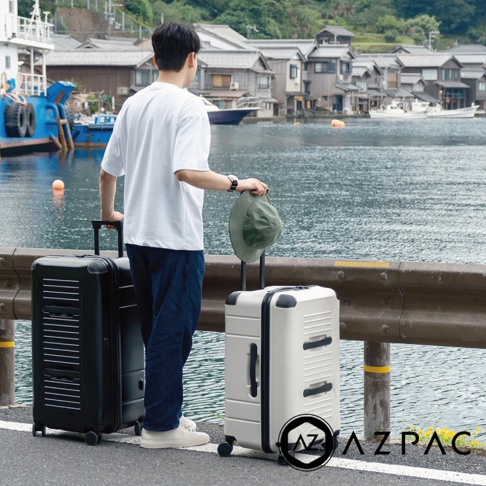 一位男士站在一條有著傳統日式建築的港口旁，身旁是兩個AZPAC Trucker系列旅行箱，一個為30吋的白色防爆拉鍊旅行箱，另一個是黑色旅行箱。
