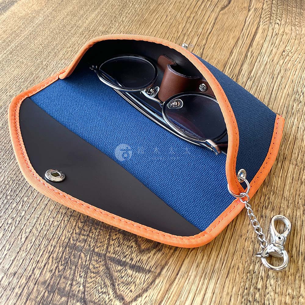 展示了犬印純綿6號帆布眼鏡收納盒，內含一副金屬框眼鏡，收納盒外緣為橘色，內襯為藍色，並帶有掛鍊和小掛鉤。