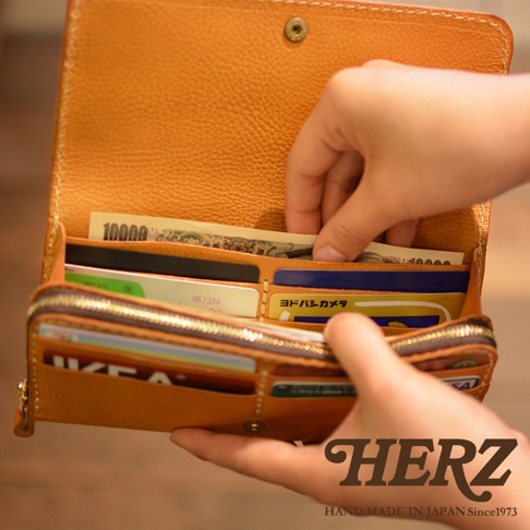 職人手作皮革長夾，呈現天然皮革紋理，顏色為溫暖的橘棕色。長夾內部分層清晰，可見多張信用卡、日幣鈔票。拉鍊設計順暢，整體質感細膩。