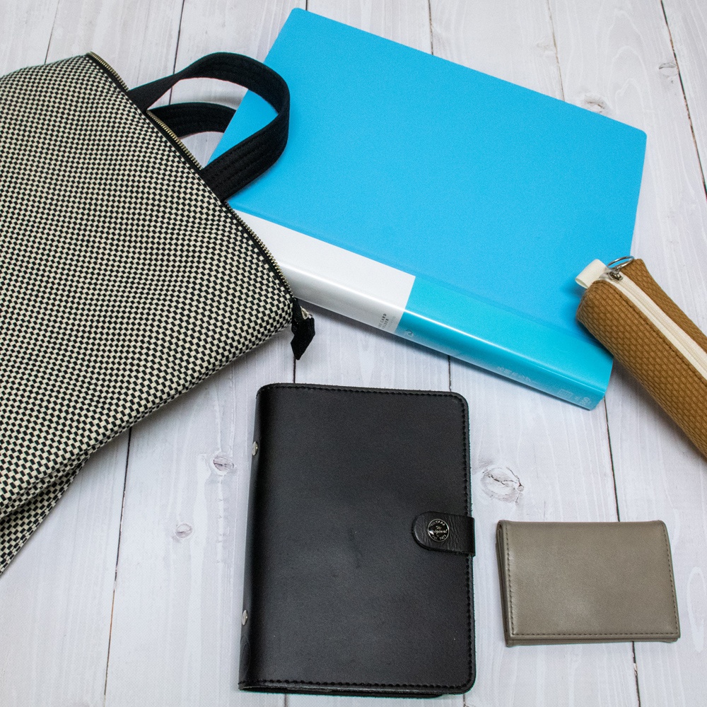 黑色皮革記事本、灰色卡夾、藍色文件夾、棕色筆袋，以及一個黑白格紋的帆布手提後背包，展示在木紋桌面上。