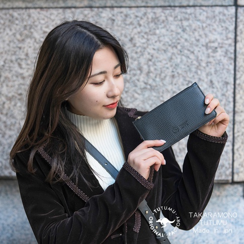身穿深棕色絲絨外套的年輕女性，展示著一款黑色荔枝紋皮革長夾，夾上印有「TUTUMU」字樣及袋鼠圖案，旁印「TAKARAMONO TUTUMU Made in jpn」。