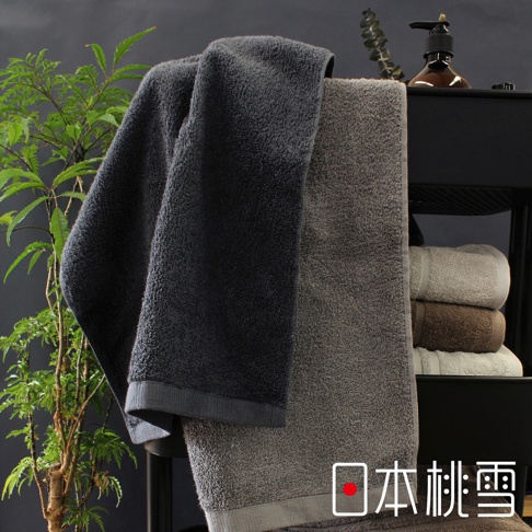 日本桃雪品牌標誌在兩條深灰色和淺灰色的厚實毛巾旁，左側有綠色植物，右側有瓶裝沐浴乳。