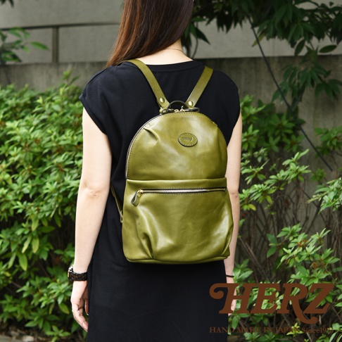 身揹橄欖綠皮革圓角後背包，有大開口設計與前方拉鍊口袋，皮革質感自然，背後有綠葉植栽。