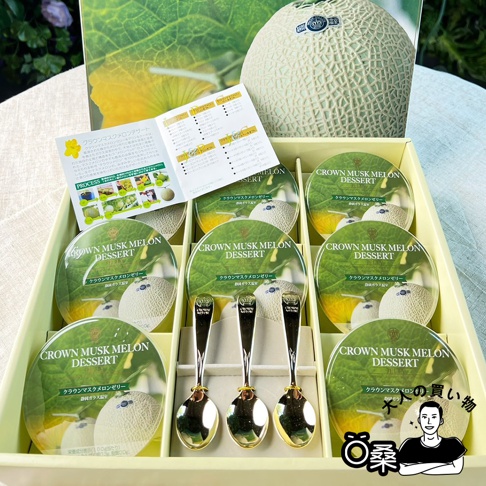 黃色禮盒內含九個圓形哈密瓜果凍，上方印有綠色哈密瓜和皇冠標誌。禮盒中央擺放著三支金柄銀質湯匙。背景為一整顆淺綠色網紋哈密瓜和產品說明書。