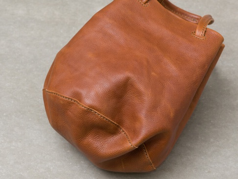 棕褐色皮革手提托特包，材質柔軟，帶有自然的紋理和縫線細節。包款呈現圓潤的輪廓，提把連接處有明顯的車縫。