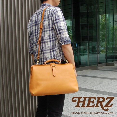 男士身揹一個淺棕色軟式皮革杜勒斯包，包包有提把和可拆卸肩帶，配戴者身穿藍白格紋襯衫。