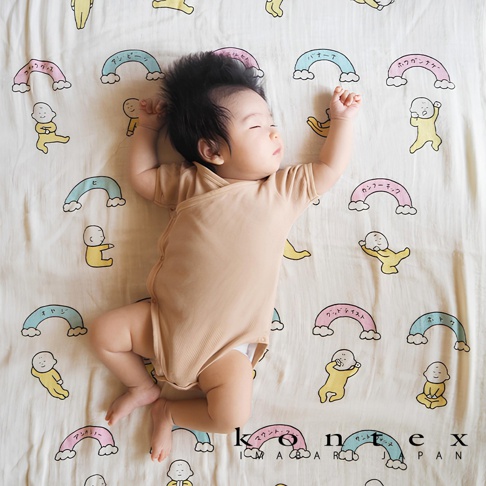 新生兒仰臥在印有彩色日文文字和可愛嬰兒圖案的淺米色紗布毯上，身上穿著淺褐色連身衣。