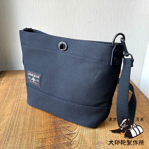 犬印鞄製作所黑色純綿帆布迷你側背包，背包上有日文品牌標籤，肩帶固定於黑色金屬扣環上。