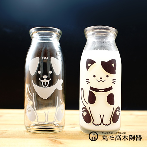 一組兩個玻璃牛奶瓶，一個是透明瓶身印有白色狗狗圖案，另一瓶裝有白色液體，瓶身印有白色和黑色貓咪圖案。