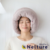 一位女士頭部被柔軟的粉色毛巾枕包覆，枕頭呈甜甜圈狀，材質類似今治毛巾，表面有細緻的紋理。枕頭一側有一個小小的紅藍色標誌。