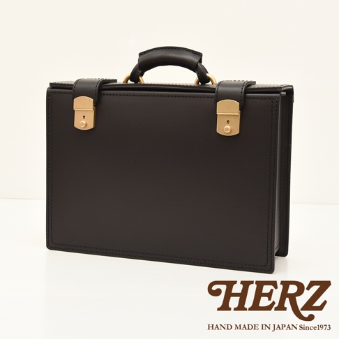 職人手作黑色皮革機師箱，設有金色鎖扣、提把與金屬配件，簡約設計，質感細緻。