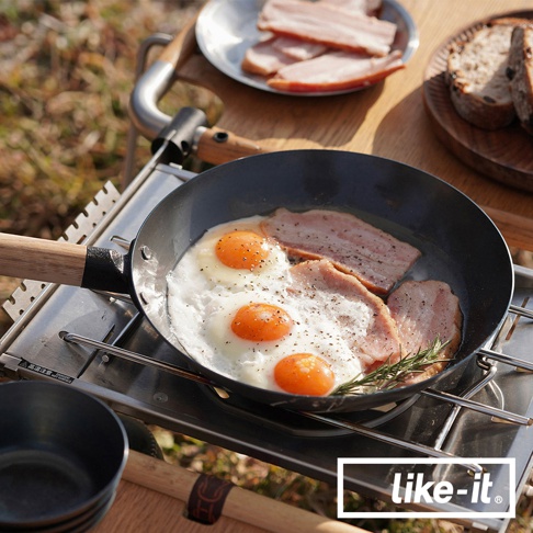 一個黑色萬能調理鐵鍋，鍋內有煎蛋和培根，鍋柄為木質，放在露營爐架上。