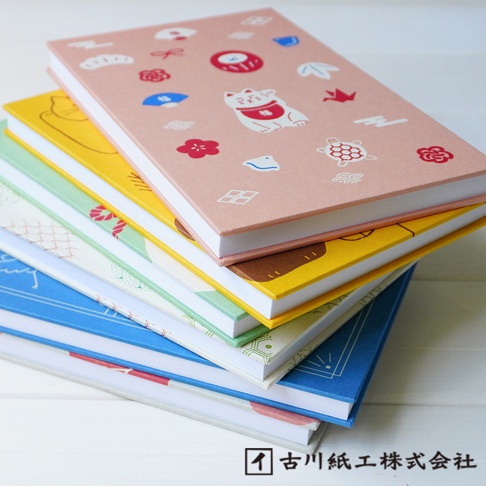 一本粉色封面的和紙御朱印帳，上面印有招福貓、花朵、烏龜、紙鶴等日式圖案。下方堆疊著黃色、綠色、藍色等多種顏色的御朱印帳。