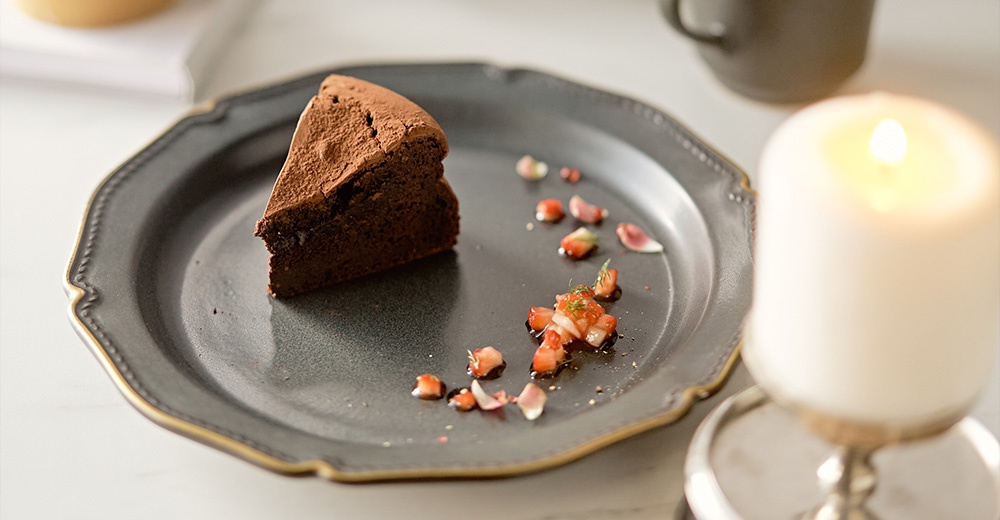 Aito Stitch 美濃燒金彩浮雕餐盤上的巧克力蛋糕，旁邊擺著點綴著草莓和香草的醬汁，以及一根燃燒的白色蠟燭。