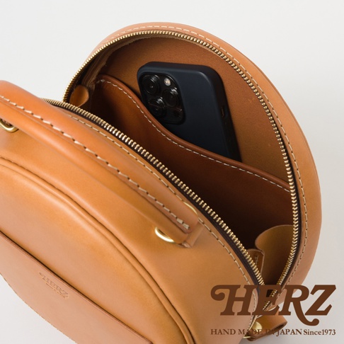 日本HERZ手工皮革圓形包，棕色皮革，內部有一個手機，上面有品牌Logo和「HAND MADE IN JAPAN Since1973」字樣。