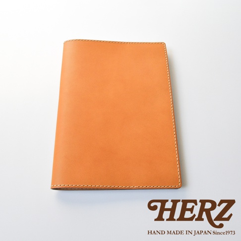 一張日本職人品牌 HERZ 手作皮革筆記本書套的產品照，採用質感溫潤的淺棕色皮革製作，邊緣有細緻的縫線工藝，呈現簡約洗練的商務風格，下方印有 HERZ 品牌名稱與手作產地字樣，背景為乾淨的純白底色。