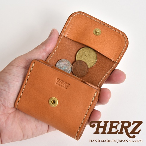 一隻手拿著一個日本HERZ職人手作的淺棕色皮革零錢包，包內裝有幾枚硬幣，皮夾表面壓印有HERZ標誌與MADE IN JAPAN字樣，整體呈現精緻的車縫細節與皮革質感。