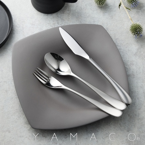 霧面不鏽鋼晚餐叉、湯匙和刀子，擺放在灰色的盤子上，背景有模糊的黑色杯子和裝飾性的植物。