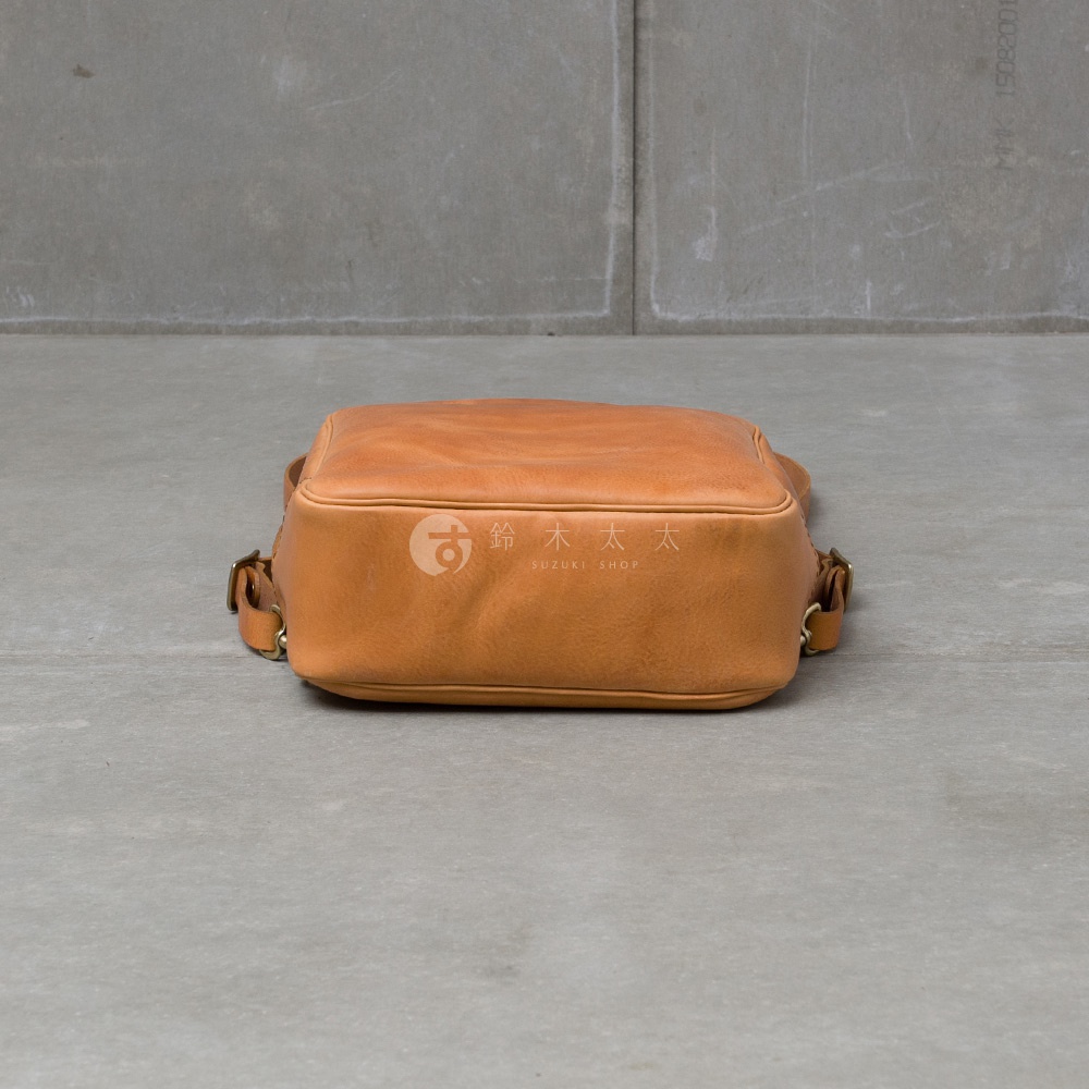 職人手作皮革Organ商務後背包M-10，淺棕色，皮革材質，包體呈現方形，置於灰色水泥地板上，背景為水泥牆面，包身有金屬扣環及提把。