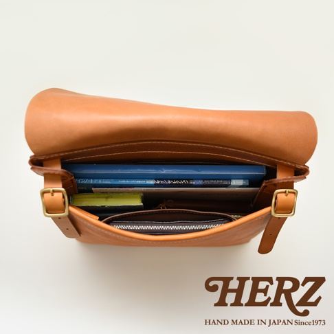 職人手作皮革經典郵差包，採用棕色牛皮製作，包包開口朝上，內有書籍、筆記本及拉鍊夾層，搭配黃銅釦環與可調節式揹帶。