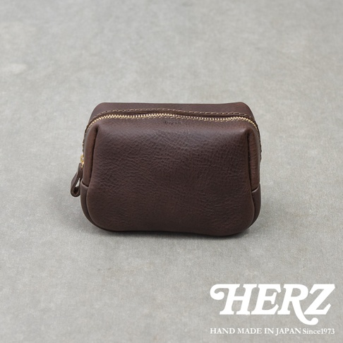 一個放置在淺灰色背景上的深咖啡色日本HERZ品牌手工皮革拉鍊收納包，皮革表面呈現細緻的自然紋理，頂部設有黃銅色拉鍊，包身線條簡約圓潤，右下角印有HERZ及其品牌創立年份的白色標誌。