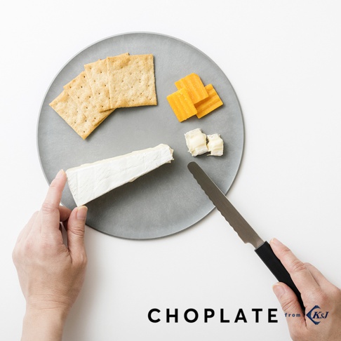 灰色圓形砧板上擺放著方形餅乾、黃色起司塊、白色軟質起司，左手拿著一塊白色起司，右手握著黑色手柄的鋸齒狀刀具。