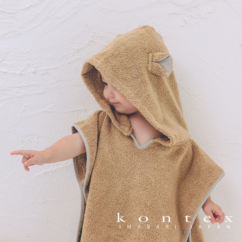 一名嬰兒穿著一件淺棕色、毛絨絨的斗篷式連帽浴巾，浴巾上有可愛的熊耳朵設計，邊緣有灰色滾邊。