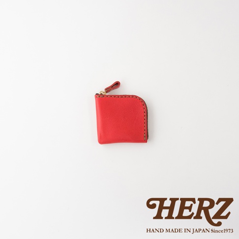 一個由日本HERZ職人手作的紅色皮革零錢包，造型精巧方正且邊角圓潤，頂部配有金屬拉鍊與皮革拉環，右側有手工縫製的細節，右下角標註有HERZ品牌字樣與日本製造資訊。