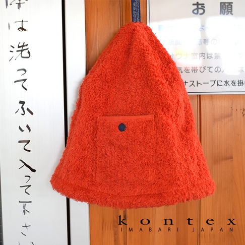 橘紅色毛巾材質的桑拿帽，帽頂有小口袋及藍色鈕扣，毛巾邊緣有流蘇設計，掛在木門旁。