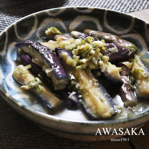 青釉繪料理盤盛裝著切塊的茄子，茄子呈現深紫色，表面帶有油光，並撒上蔥花與其他調味料。盤子邊緣飾有藍白相間的紋飾。