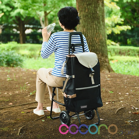 一位短髮女子坐在公園樹下休息，正在飲用瓶裝水。她背後放置了一款 COCORO 多功能摺疊購物車，這款購物車採黑米色帆布設計，具備摺疊座椅功能，下方裝有輪子，整體呈現出戶外休閒與便利購物的機能感。