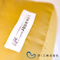 一條白色標籤縫在黃色布料上，標籤上有黑色字樣「life is a great JOURNEY」，下方有日文標誌及「まくら株式会社」。