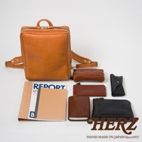 展示職人手作皮革軟皮商務後背包，搭配各式棕色、黑色皮革配件，包含長夾、化妝包、筆袋及筆記本，整體呈現在白色背景上。