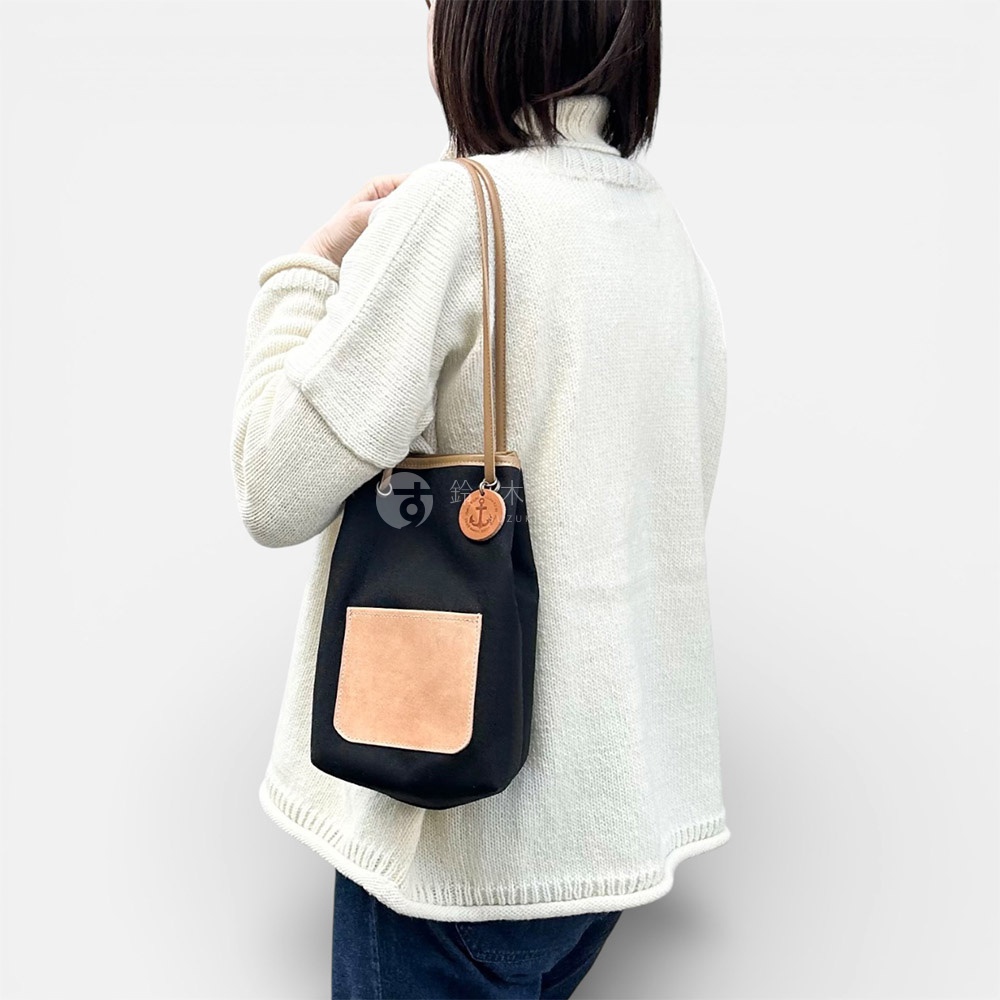 一名女子背對鏡頭，身穿白色高領毛衣，肩揹一個黑色束口袋包，包上裝飾著一個咖啡色皮革製的船錨吊飾。