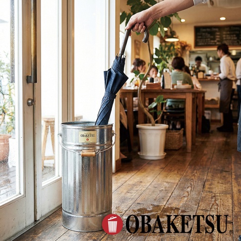 OBAKETSU 手工鍍鋅鋼製傘桶，內側放著一把深藍色雨傘，放在木地板上，背景是咖啡館的座位區。