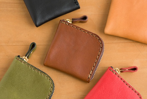 多款職人手作皮革收納零錢包，包含棕色、綠色、紅色、黑色及橘色，皆為 L 形拉鍊設計，搭配金色拉鍊頭與皮革拉環，邊緣縫線清晰可見。