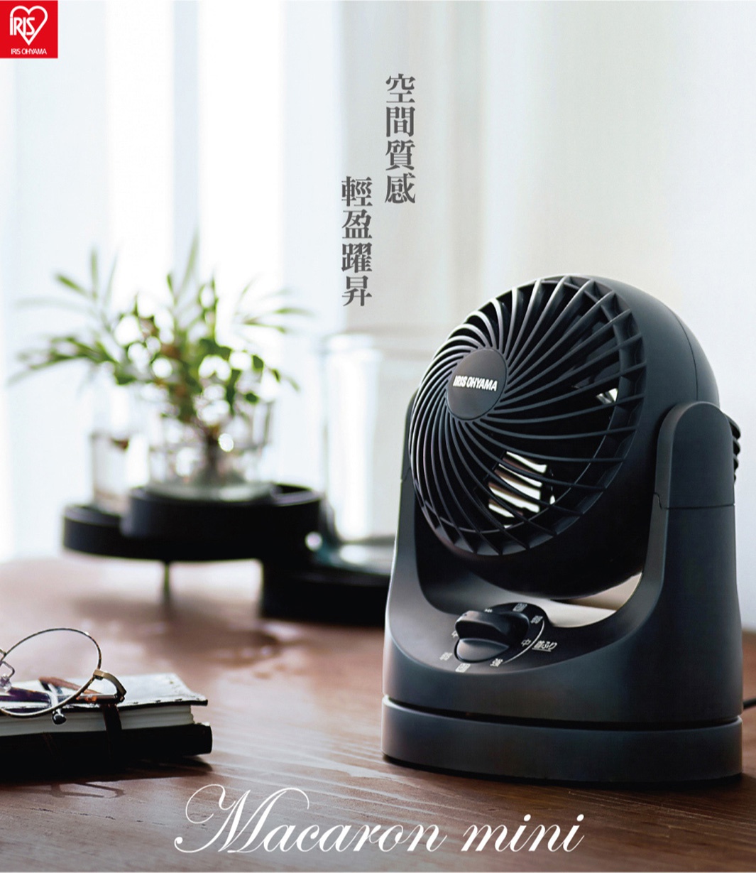 黑色迷你型空氣循環扇，具有圓潤的機身和可調節風速的旋鈕，置於木質桌面上，旁有一疊筆記本和眼鏡。