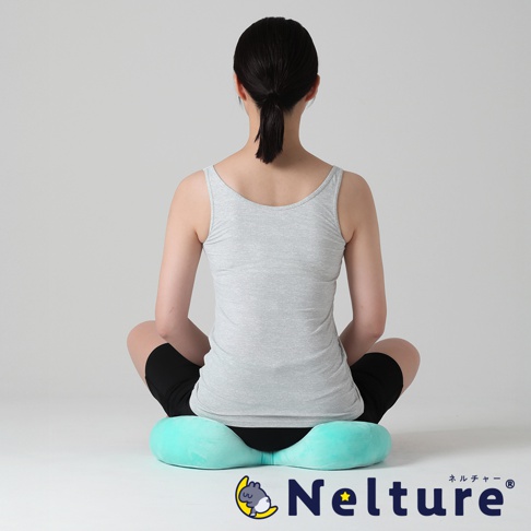 一位女性從後方視角坐著，她穿著灰色細肩帶背心和黑色短褲，坐在一個土耳其藍色的瑜珈感舒壓美姿矯正坐墊上，坐墊的下方可以看到品牌名稱 Nelture。