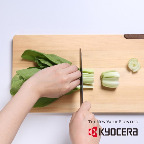 左手持綠色青江菜，右手使用黑色刀刃的陶瓷刀，在木質砧板上切細，砧板上另有切好的青江菜段。