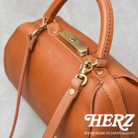 職人手作皮革Organ兩用口金手提包，採用光滑的棕色皮革，配有金色金屬口金鎖扣、提把和可拆卸肩帶。包身呈現圓筒狀，皮革縫線清晰可見。