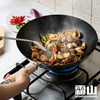 日本製木柄窒化鐵炒鍋，鍋內有蛤蜊、辣椒和蔥段正在烹調，鍋身呈黑色，鍋柄為木製，放置於瓦斯爐上。