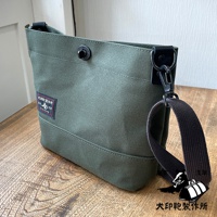 軍綠色純棉帆布迷你側背包，配有黑色織帶和金屬扣環。包身有縫線裝飾，左側印有紅色標籤，頂部有黑色按扣。